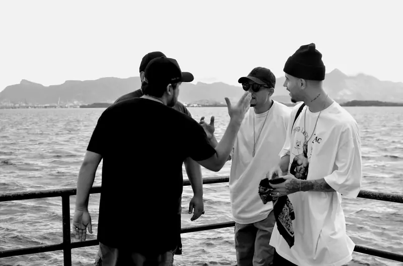Integrantes do 1DV conversando num píer na Baía de Guanabara, com as montanhas do Rio ao fundo